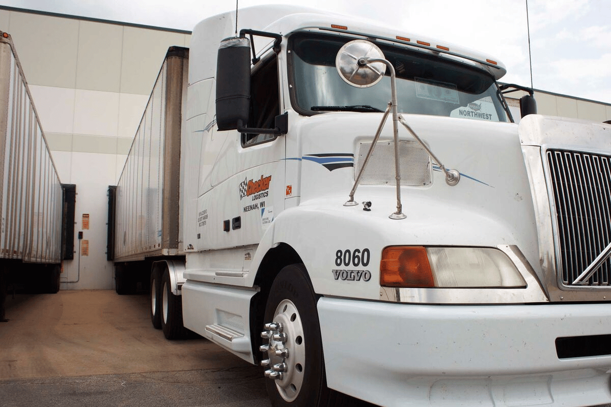 White semi-truck with a trailer parked at a loading dock of a warehouse, marked with company logos.