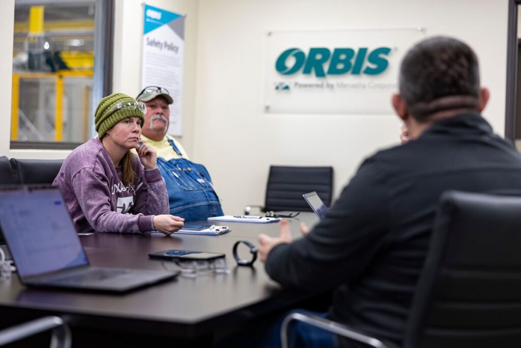 Three people sitting at a conference table during a meeting in an office. Two laptops and a phone are on the table. A sign on the wall reads "ORBIS.